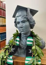 A bust of a young man wearing a graduation cap, adorned with a green and white scarf and a garland of leaves, displayed in a library setting.