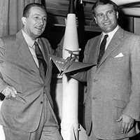Two men in business suits standing next to a model rocket, with one holding a small airplane model.