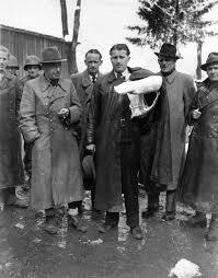 A historical black and white photograph of a group of men standing outdoors, wearing various styles of hats and coats. One man in the center holds a rolled-up document, while others observe him.
