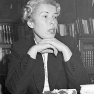 A woman with light-colored hair styled in soft curls, wearing a dark blazer, sitting at a table with her hands clasped, against a background of bookshelves.