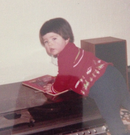 A toddler with a brown bowl haircut is leaning on a 1970s hifi stereo resting a book on top.