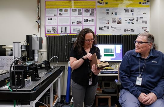 A medium wide shot of a man and a woman in a lab with several pieces of equipment along the wall. She is standing in center of the room, looking at a book, and he is seated on the right, looking over at her.