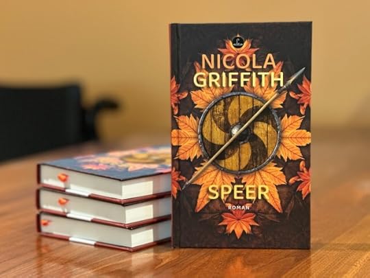 A table on which lie 3 copies of a novel, SPEER by Nicola Griffith, next to which stands a single copy facing the camera. The cover in autumnal hues shows a shield lying on leaves, over which lies a spear