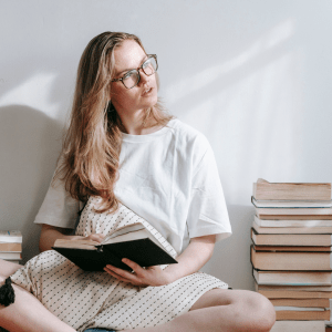Woman sitting cross-legged on floor, reading from a stack of books.
