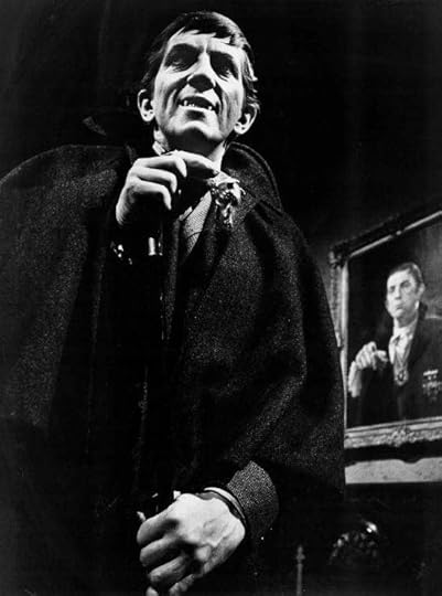 Black and white image of a man in a cloak holding a cane, smiling while looking at the camera; a portrait of another man is visible in the background.