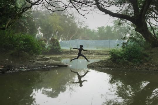 Image: a child leaps from one bank to another over a narrowing in a creek.