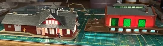 Two n-scale railroad buildings on a workbench, both have bright light spilling out of their windows.
