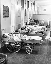 A black and white photograph of a hospital corridor with multiple patients resting on beds, some on wheelchairs, highlighting a clinical environment.