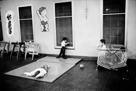 Black and white photo of three children in a room, with two near windows and one lying on a play mat.