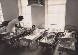 A historical black and white image of a nurse attending to infants in cribs inside a care facility.
