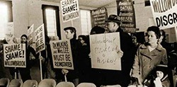 Black and white photograph of a group of protesters holding signs with messages like 'Shame!' and 'Disgrace!' inside a hall.