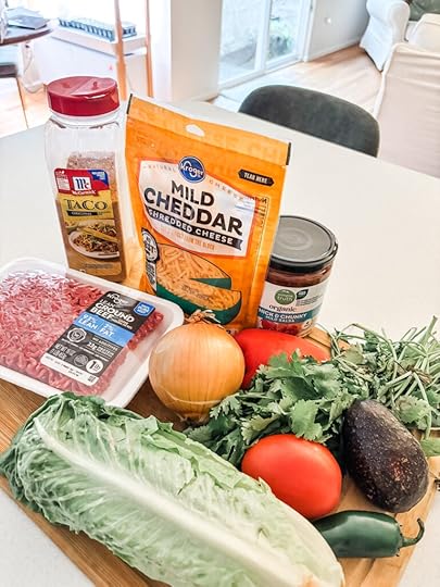 Ingredients for a low-carb taco board laid out on a counter, including ground beef, McCormick taco seasoning, romaine lettuce, avocado, roma tomatoes, jalapeño, shredded cheese, salsa, and fresh cilantro