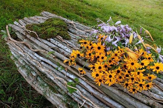 Photo of a beautiful coffin fashioned out of tree branches. The coffin lays on the grass and has yellow and purple flowers on top of it
