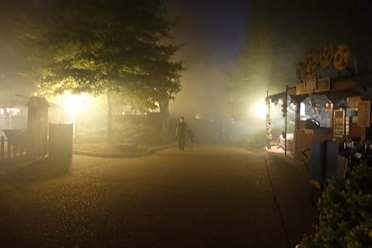 A foggy pathway at night in a park, illuminated by soft lights, with trees on either side and two figures walking in the distance.