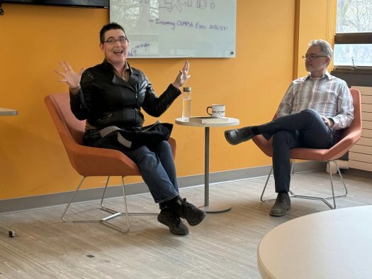 Ada Hoffmann is wearing short hair, glasses, a leather jacket, jeans, and black running shoes. They are sitting in a red chair in front of a yellow wall, waving their hands in an expressive gesture. They look happy and excited. Beside them is a small table with a water bottle, copy cup, and book. On the other side of the table a man with gray hair sits. He is wearing a striped shirt, jeans, and brown shoes. He is the event host.