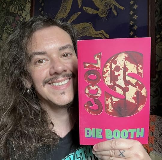 Image of author Die Booth holding up a copy of Cool S and smiling. He is a middle aged white man with dark eyes and facial hair and long curly dark hair. Cool S has a hot pink cover with author name in green and the title 'cut out' to reveal a bloody cool s behind.
