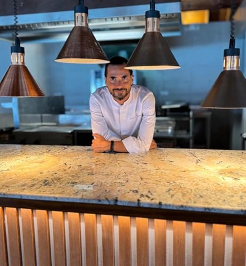 A chef in a white uniform leans on a granite countertop in a modern kitchen, illuminated by hanging industrial-style lights.