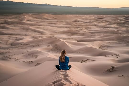 A woman with her back to us sits cross-legged among barren sand dunes, looking out.