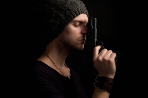 Man in beanie holding a gun pointing upward and resting against his face, eyes closed, looking regretful.