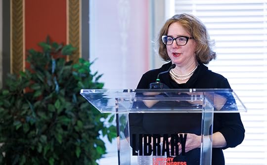 Medium shot of a woman speaking at a clear podium. she is wearing a dark suit and glasses. The room is formal, with well-lit windows behind her. 