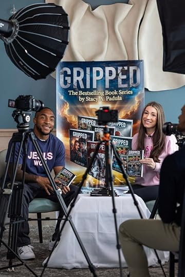 A man and a woman sitting at a table holding books from the 'Gripped' series by Stacy A. Padula, with cameras set up for an interview. A promotional banner for the book series is visible in the background.
