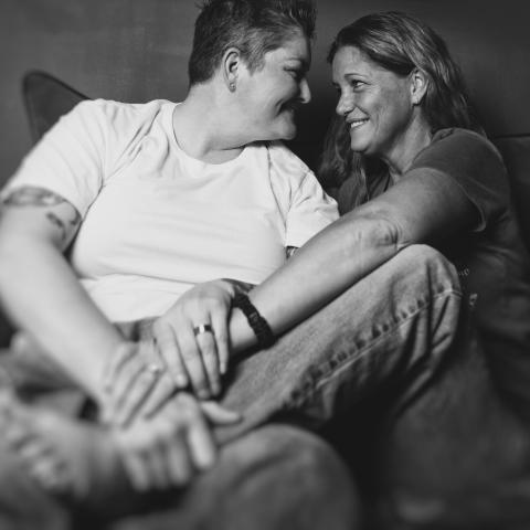 A black-and-white photo of a couple, limbs intertwined, on a couch. They're gazing into each other's eyes. 