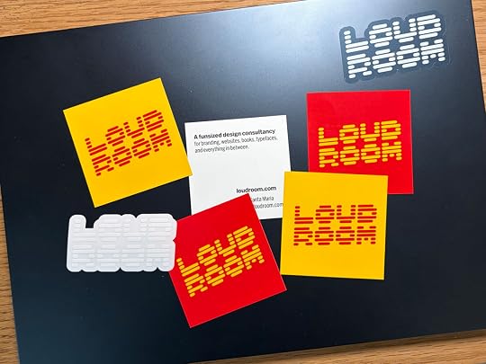 Overhead photo of a closed laptop with various yellow/red and clear Loud Room stickers strewn around. A square sticker back in the middle has black text on a white background: A funsized design consultancy for branding, websites, books, typefaces, and everything in between.