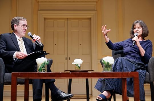 A man and woman, seated onstate in chairs behind a coffee table, are smiling as the woman speaks and raises her right hand to make a point.