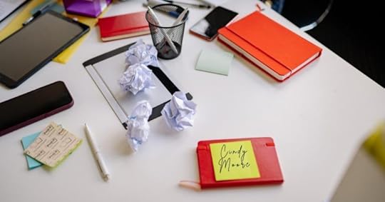Promotional image for a writing or publishing announcement. The photo shows a cluttered white desk covered with notebooks, a tablet, smartphones, sticky notes, pens, and several crumpled sheets of paper, suggesting a brainstorming or writing process. In the foreground, a yellow sticky note attached to a red notebook bears the handwritten name “Cindy Moore.” The scattered materials and discarded drafts convey the creative challenges of developing a new story, character, or book series, while the clean workspace and bright lighting evoke inspiration, planning, and the early stages of a writing project.