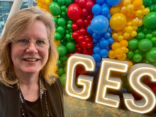 A selfie of Jen in front of a balloon wall in green, red, blue, and yellow, with a lit up sign that says G-E-S.