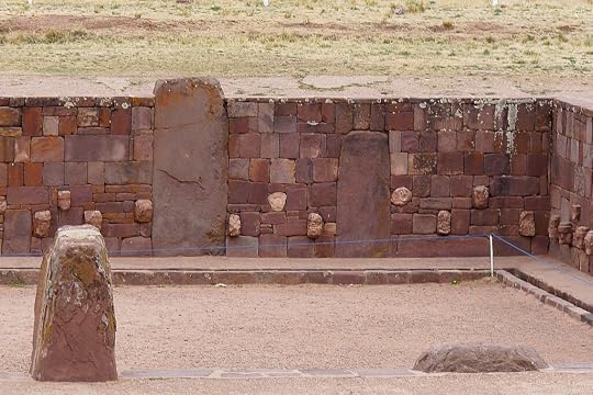 An archaeological site featuring a stone wall made of intricately cut reddish-brown stones, with carved figures embedded within the wall. In the foreground, a large stone monument is visible.