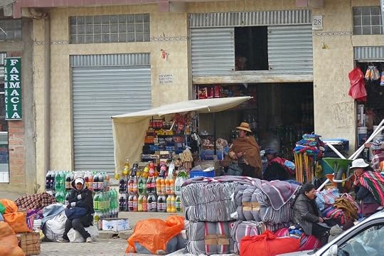 A street market scene featuring a small shop with a variety of products displayed outside, including soft drink bottles, and several people sitting on the ground near colourful blankets and bags.