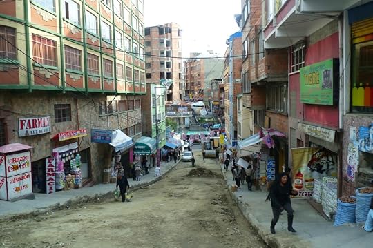 View of a sloped street in a vibrant urban area with various shops and people walking. Buildings line the street, featuring signs in Spanish, while pedestrians navigate the uneven pavement.