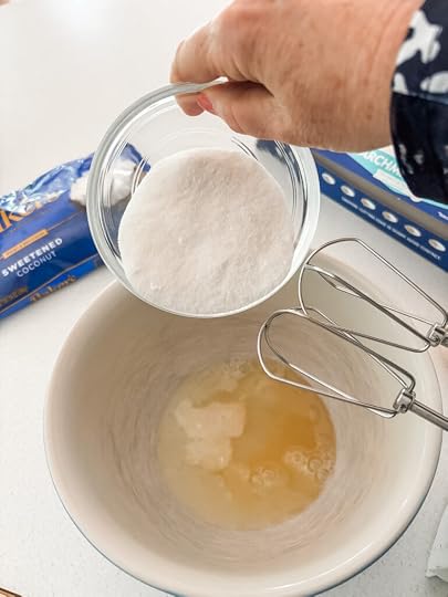 Whisking egg whites, sugar, and vanilla together in a glass bowl before adding shredded coconut