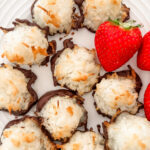 Close-up of a chocolate-dipped coconut macaroon showing its golden toasted exterior and dark chocolate base with strawberries