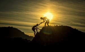 Silhouette of one man helping another man to reach the top of a mountain.