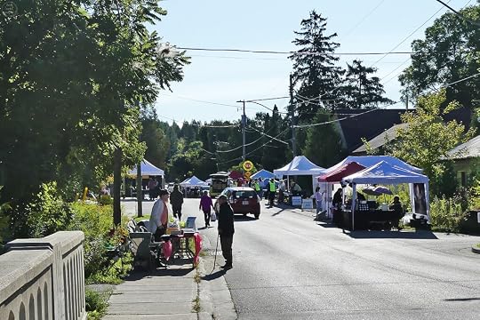 A street scene featuring several vendor tents set up for a market day, with people walking and shopping, surrounded by trees and buildings.