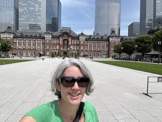 A selfie of me in sunglasses and a green shirt standing in front of Tokyo Station, a large red brick building with white stone accents that looks like a large English manor house.