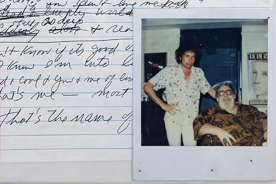 A color snapshot of two men, one standing and one seated, both looking at the camera and smiling. It is next to a notebook page with scribbled song lyrics.