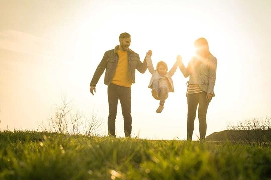 Happy parents walking in nature swinging their daughter between them, backlit by sun.