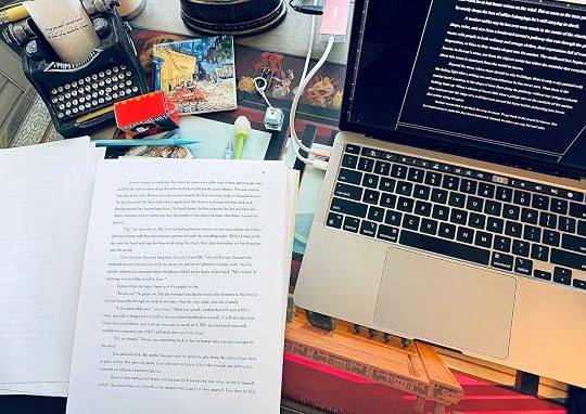 photo of my writing desk with my computer, printed pages, a typewriter decoration, a french coaster, and a music box