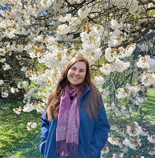 smiling brunette woman in a blue coat and purple scarf standing before white blossom