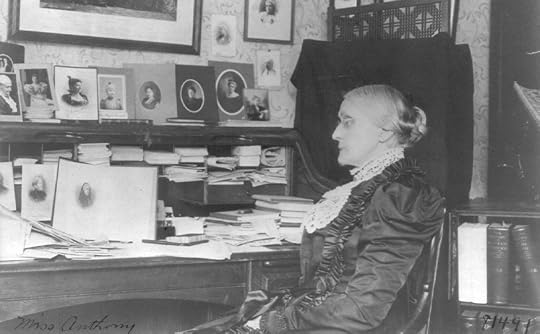 A woman with white hair pulled back into a severe bun, seated at rolltop desk, opened to show a scattered papers and several framed photographs. 