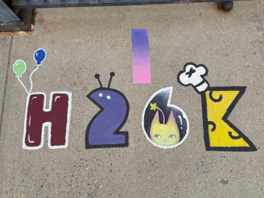 Photograph of some graffiti painted onto a concrete sidewalk, spelling H 2 6 K. The ���H��� has two balloons tied to it, the ���2��� is stylized to look like a little alien, the ���6��� has a little yellow cat inset into it, and the ���K��� has a chef���s hat.