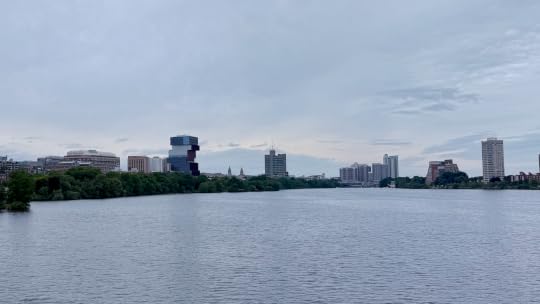Photograph of the Charles River, looking west toward Allston and Cambridge. Some buildings are visible on the horizon, and the banks of the river are covered with green leafy trees.