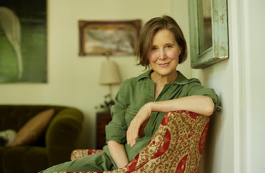 A half portrait of Ann Patchett, who is seated sideways in an upholstered chair in a living room, smiling at the camera. 
