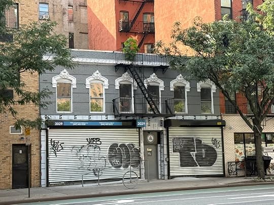 A street view of a gray building with a decorative façade, featuring multiple windows and a fire escape, with graffiti on the metal storefront shutters. Nearby, there are trees and other buildings in the background.