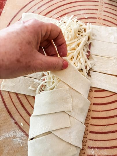Hands folding cut strips of puff pastry over a ham and cheese filling in a faux-braid pattern on a parchment-lined baking sheet