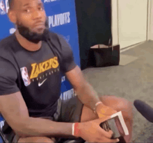 a man wearing a black lakers shirt sits in front of a blue wall