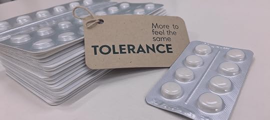 Stacks of silver medication blister packs surround a brown paper tag reading “TOLERANCE,” with the smaller phrase “More to feel the same.”